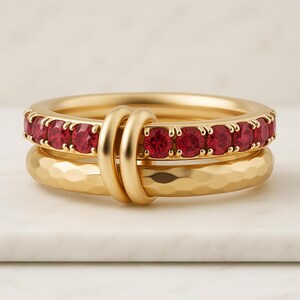May include: Three gold rings. The top ring is adorned with a row of red gemstones. The middle ring is connected to the top ring by two gold loops. The bottom ring has a hammered texture. The rings are stacked together.