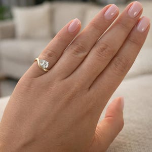 May include: A gold ring with a pear-shaped diamond. The ring's band curves around the diamond, creating a unique and elegant design. The ring is shown on a hand with polished nails.