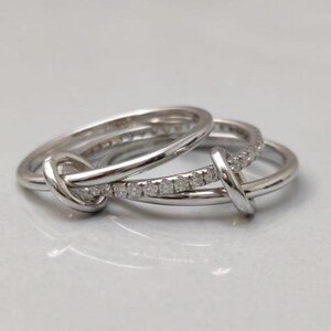 May include: Three silver rings interlock in a unique design. One ring features a row of small, sparkling stones. The rings are intertwined, creating a knot-like appearance. The rings are set against a soft, neutral background.