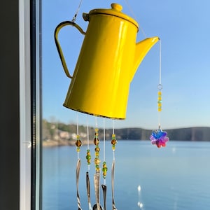 May include: A whimsical wind chime crafted from a yellow enamel teapot, suspended by a window. Silver spoons hang below, adorned with beads in shades of green and brown. A colorful flower-shaped charm adds a playful touch.