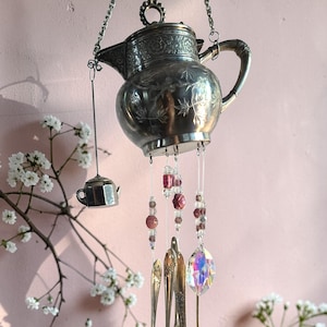 May include: A decorative wind chime featuring a silver teapot, spoons, and a crystal prism. The teapot is suspended from a chain and adorned with floral patterns. The chime includes red beads and vintage spoons, creating a unique, handcrafted home decor item.