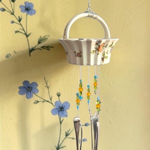 May include: A hanging ceramic basket wind chime with floral accents and colorful beads. Three silver spoons hang below the basket. The background is a yellow wall with blue floral patterns.