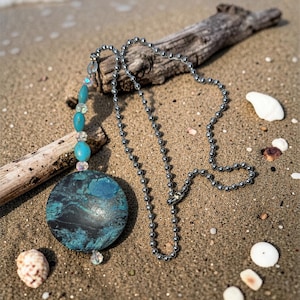 May include: A necklace with a large, round pendant featuring a blue and black marbled design. The pendant is connected to a silver ball chain. The necklace is displayed on a sandy beach with seashells and driftwood.