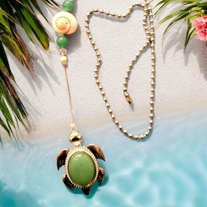 May include: A gold-tone turtle charm with a green gemstone pendant hangs from a silver chain with green and white beads. The chain is attached to a white ceiling fan pull.