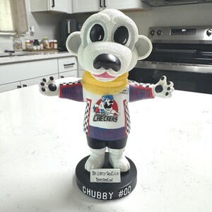 May include: A white bear bobblehead figure wearing a Charlotte Checkers hockey jersey with red and blue sleeves, a yellow scarf, and black shorts. The base features the text CHUBBY #00 and a plaque reading Dr. Larry Seitlin Team Dentist.