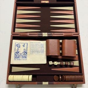 May include: A brown leather backgammon set featuring a dark brown felt interior with cream and brown triangular points. The set includes two brown leather dice cups, two white dice, two brown dice, a doubling cube, and cream and brown checkers.