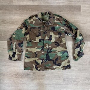 May include: A long-sleeve field jacket featuring a woodland camouflage pattern in shades of olive green, brown, and black. The jacket includes a fold-down collar, a front button closure, two chest pockets with flaps, and two lower pockets with flaps.
