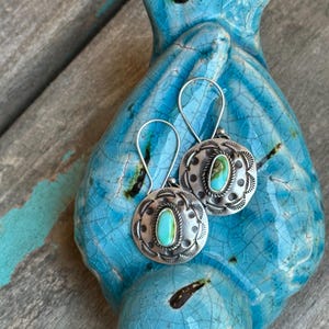 May include: A pair of silver earrings with turquoise stones set in a circular design. The earrings are hanging from a blue ceramic surface.