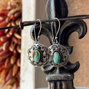 May include: A pair of silver earrings with turquoise stones set in a circular design. The earrings are hanging from a metal stand.
