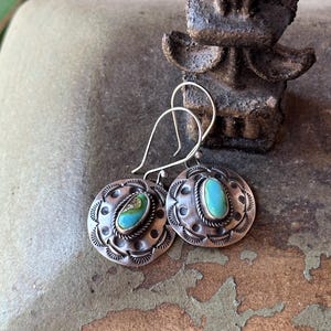 May include: A pair of silver earrings with turquoise stones set in a circular design. The earrings have a simple wire hook closure.