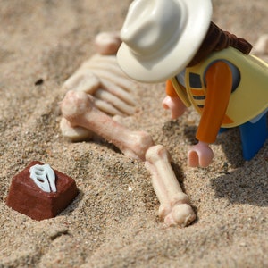 Puede incluir: Una figura de juguete de plástico de un explorador que lleva un sombrero marrón y un chaleco amarillo, arrodillado junto a un esqueleto de plástico blanco sobre una superficie arenosa. Un pequeño ladrillo marrón con un hueso de plástico blanco en la parte superior está en primer plano.