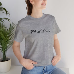 May include: A heather gray t-shirt with the text "PH.inished" printed in black. The shirt is a crew neck style and appears to be made of a soft material. The person is wearing blue jeans.