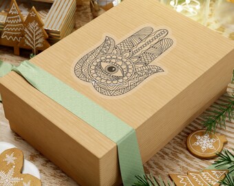 Può includere: Una scatola regalo marrone con un nastro verde e un adesivo di mano di hamsa in bianco e nero con un occhio al centro. La mano di hamsa è un simbolo di protezione e buona fortuna.