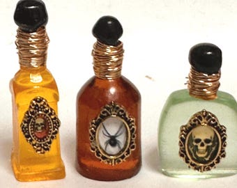 May include: Three small glass bottles with gold wire around the necks and decorative gold frames with spooky images inside. The bottles are filled with yellow, amber, and green liquids.