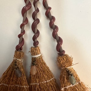 May include: Three miniature brooms with brown twisted handles and natural fiber bristles. Each broom has a decorative charm with a gemstone hanging from it.