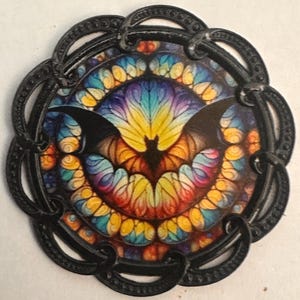 May include: A round, black metal pendant with a colorful stained-glass design. The design features a bat silhouette in the center, surrounded by a vibrant array of colors including yellow, orange, blue, and purple. The pendant has an ornate, looped border.