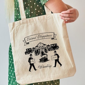 May include: A beige canvas tote bag with black graphic illustrations. The design includes a banner with the words "Eternal Happiness", vintage cars, people, and the word "Wembley". The bag has two handles.