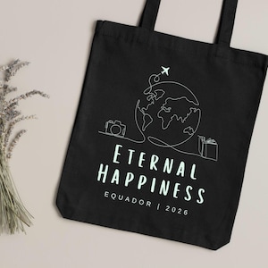 May include: Black tote bag with a white line drawing of a globe, airplane, camera, and gift. The words "Eternal Happiness" and "Equador | 2026" are printed in a light green color. A bundle of dried lavender is on the left.