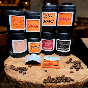 May include: A collection of eight black coffee canisters with various labels, including "Dark Roast", "Espresso", "Medium Roast", "Coffee", "Decaf", and "Southern Weather". The canisters are arranged in two rows of four on a wooden table with coffee beans scattered around them.