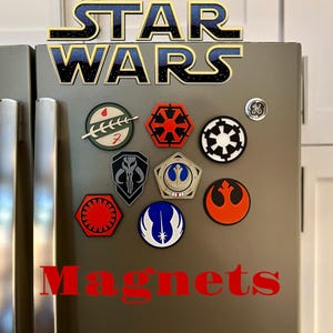 May include: A set of nine Star Wars themed magnets featuring various symbols and characters from the franchise. The magnets are attached to a stainless steel refrigerator.
