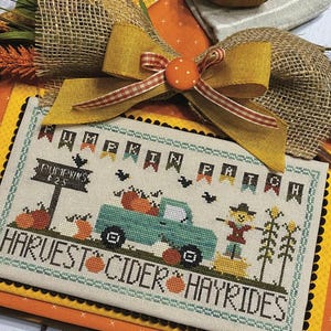Könnte beinhalten: Ein Kreuzstich-Design mit dem Text "Pumpkin Patch" und "Harvest Cider Hayrides". Das Design zeigt einen tealfarbenen Truck mit Kürbissen, eine Vogelscheuche und den Text "Primrose Cottage Stitches". Über dem Design befindet sich eine Schleife aus Jute mit gelbem Band und orangefarbenen Akzenten.