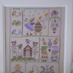 以下が含まれることがあります： パステルカラーの春らしい風景が描かれたクロスステッチ刺繍の額縁。ウサギ、鳥、花、小さな家などが描かれています。フレームの下部には「Cuore e Batticuore」というテキストがあります。