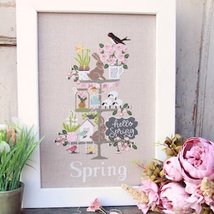Puede incluir: Un diseño de bordado de punto de cruz con un árbol de temática primaveral con pájaros, conejos, flores y un letrero que dice "hello spring". El diseño está enmarcado en blanco y la palabra "Spring" está bordada debajo del árbol.