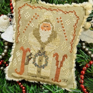 May include: A beige fabric ornament with a white, brown, and green embroidered image of Santa Claus holding a wreath. The ornament has a brown fringe border and the word "JOY" is embroidered in orange on the front.