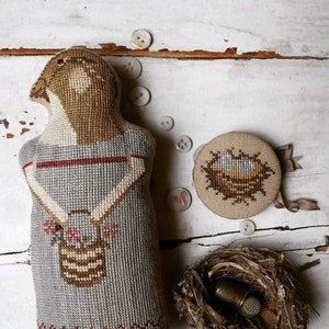 May include: A handmade, cross-stitched doll with a bird-like head, wearing a gray dress with a basket of flowers. A round, cross-stitched pincushion with a nest design and a real bird's nest with thimbles are also present. The items are on a white, distressed wooden surface.