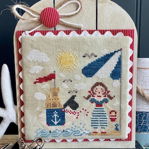 May include: A framed cross-stitch depicting a beach scene with a girl, a sandcastle, and an umbrella. The artwork features a red and white scalloped border, a wooden frame, and a decorative bow. The color palette includes blue, red, yellow, and beige.