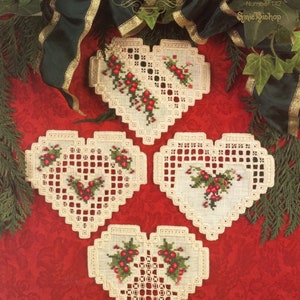 May include: Four heart-shaped, off-white cross-stitch coasters with red berries and green leaves. The coasters are arranged on a red patterned background, with text that reads "Christmas In My Heart".