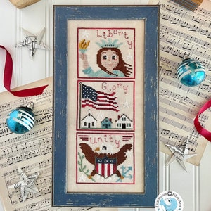 May include: A framed cross-stitch sampler with a blue frame. The sampler is divided into three sections. The top section features a woman holding a torch with the word "Liberty" above her. The middle section features a red, white, and blue American flag with the word "Glory" above it. The bottom section features an eagle with the word "Unity" above it. The sampler is surrounded by sheet music and red ribbon.