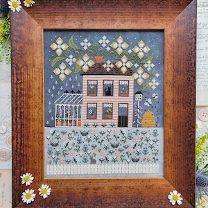 May include: A cross-stitch embroidery of a pink house with a green greenhouse, surrounded by a garden of flowers. The house has a black cat in one of the windows and a beehive in the yard. The background is a blue sky with white stars.