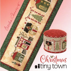 Puede incluir: Un patrón de punto de cruz con un diseño de pequeño pueblo navideño. La pieza larga y rectangular muestra casas y árboles detallados en rojo, verde y beige. También se ve un pequeño alfiletero redondo con un diseño a juego.