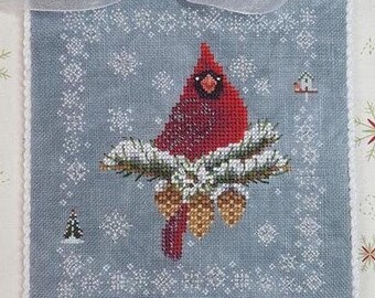 May include: A square, cross-stitched Christmas ornament featuring a red cardinal perched on a snow-covered pine branch. The bird is set against a light blue background with snowflake and pine tree accents. A white ribbon is attached for hanging.