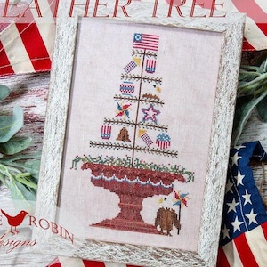 May include: A framed cross-stitch design of a "Freedom Feather Tree" with red, white, and blue patriotic decorations. The tree is in a red pot with a white rim and is surrounded by green foliage. The design is by Little Robin Designs.