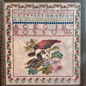 May include: A framed cross-stitch embroidery with the word "Beloved" at the top. The embroidery features a red and brown bird, a purple bird, and a floral design. The border of the embroidery is a red and green vine with small red berries.