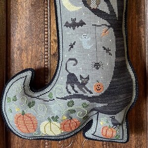 May include: A Halloween-themed, cross-stitched boot-shaped decoration. The boot features a black cat, pumpkins, bats, a ghost, and a tree with an owl. An owl decoration and fall leaves are attached to the top. The color palette includes gray, black, orange, and yellow.