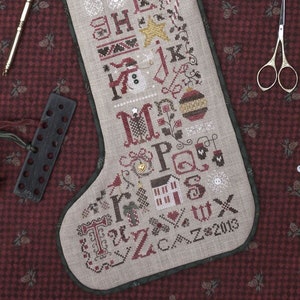 May include: A beige stocking with a green border is embroidered with the alphabet, a snowman, a Christmas tree, and the name "Cynthia". The stocking is on a red and white patterned fabric.