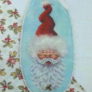 May include: Oval Christmas ornament featuring a cross-stitched Santa Claus face with a red hat, white beard, and gold-rimmed glasses. The ornament is trimmed with a white rope and a red and green ribbon with small bells. The background is light blue.