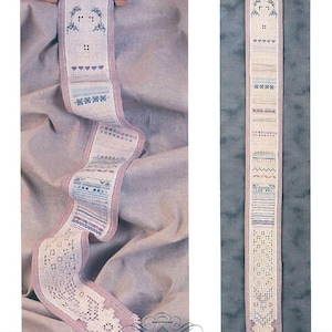 May include: An embroidered textile with a wooden hanger, featuring cross-stitch patterns. The design incorporates hearts, houses, and geometric shapes in blue, pink, and white. The bottom edge has a lace trim. The text "CROSS 'N PATCH" is visible. The textile is long and narrow.