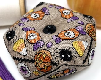May include: A beige, octagonal pin cushion with a black button in the center. The cushion is embroidered with Halloween-themed designs, including candy, spiders, pumpkins, and candy corn. A pumpkin and spools of thread are in the background.