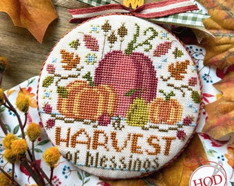 Op de afbeelding: Een ronde, geborduurde ornament met een herfstthema. Het ontwerp toont pompoenen in oranje, groen en rood, met de tekst "HARVEST of BLESSINGS". Het ornament is versierd met een ruitjeslint en herfstbladeren.