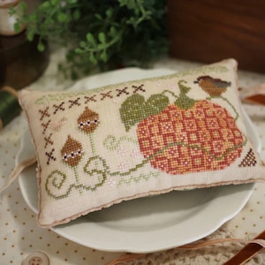 May include: A small, cream-coloured pillow with a cross-stitch design featuring a pumpkin, leaves, and a bird. The pillow is on a white plate with a brown ribbon.