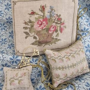 May include: A selection of cross-stitch items with floral patterns. Includes a large square piece with a basket of flowers, a small square pincushion, a rectangular pillow, and gold scissors. The items are on a blue floral patterned fabric.