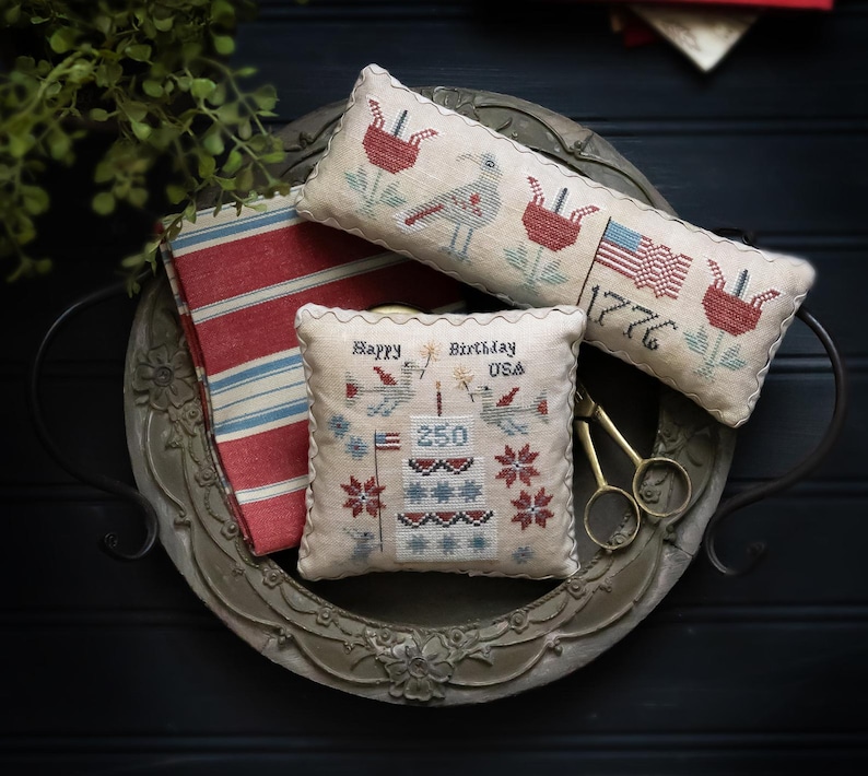 K&ouml;nnte beinhalten: Eine dekorative Anordnung mit Kreuzstichkissen im patriotischen Stil, die V&ouml;gel, Blumen und die amerikanische Flagge zeigen. Die Kissen tragen die Aufschriften "Happy Birthday USA" und "1776". Ein gestreifter Stoff und ein dekoratives Metalltablett erg&auml;nzen das Arrangement.