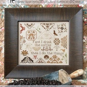 May include: A framed cross-stitch sampler with the text "First I drink the coffee then I do the things." The sampler is in a dark wood frame. Coffee beans and a scoop are in the foreground. The text "Coffee Quaker" is at the bottom.