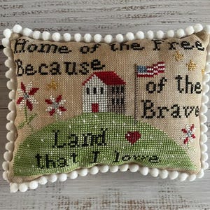 May include: A rectangular, beige decorative pillow with a white pom-pom trim. The pillow features cross-stitch embroidery with the text "Home of the Free Because of the Brave Land that I love." The design includes a house, flag, and flowers.