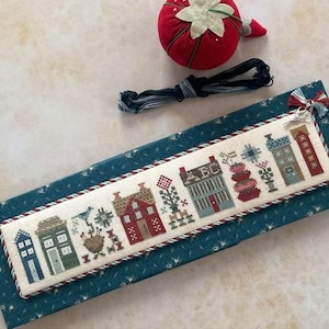 May include: A cross-stitch sampler featuring a row of colorful houses, a blue bird, and floral elements. The sampler is framed with a red and white rope trim and rests on a blue patterned surface. A red strawberry pin cushion and embroidery floss are also present.