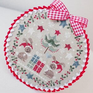 May include: A round, embroidered Christmas ornament with a red and white gingham bow. The ornament features a cross-stitch design of bunnies in Santa hats, gifts, stars, and a Christmas tree, with a red and white pom-pom trim.